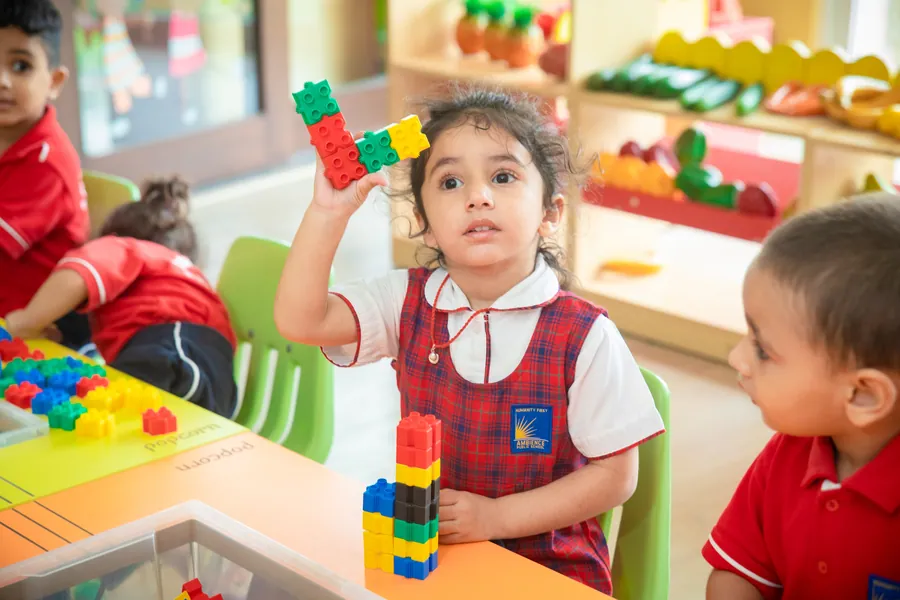 A nursery girl holding up a tower of interlocking blocks, classmates either side