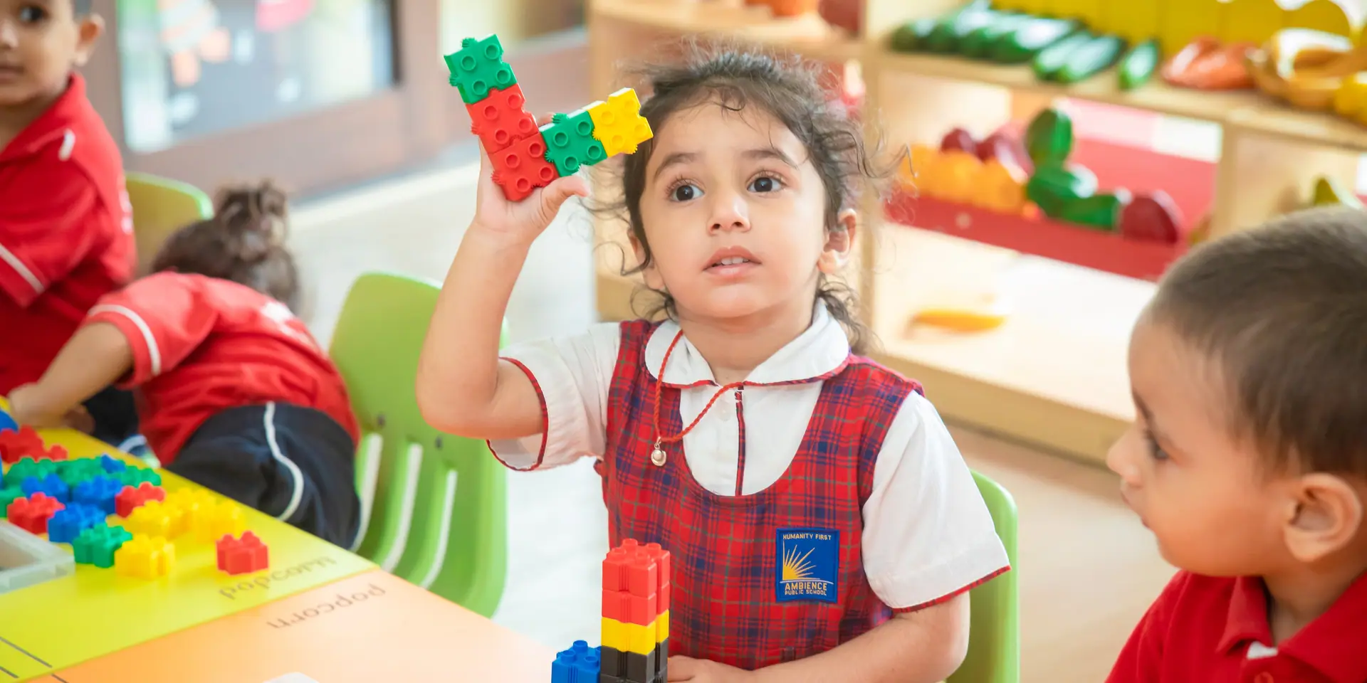 A nursery girl holding up a tower of interlocking blocks, classmates either side