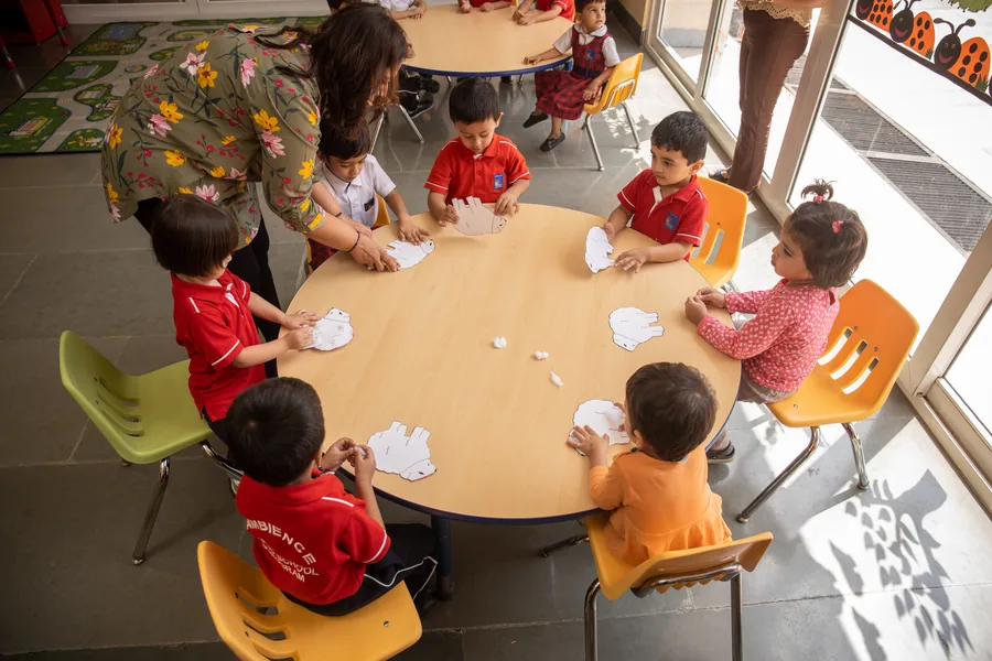 An overhead view of a teacher and nursery children doing paper craft round a table in window light