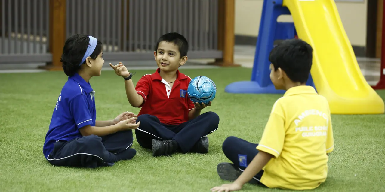 Three boys in blue, red and yellow house shirts sitting on the turf passing a ball