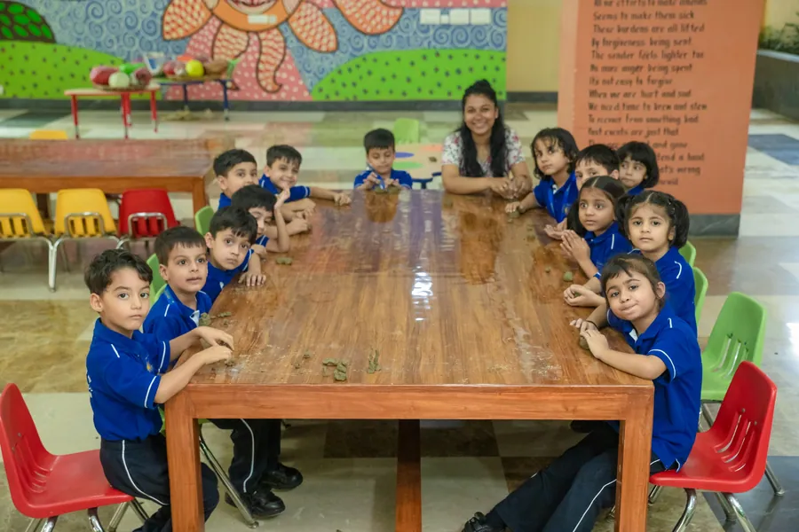 Nursery children and their teacher round a long wooden table beneath a painted flower mural