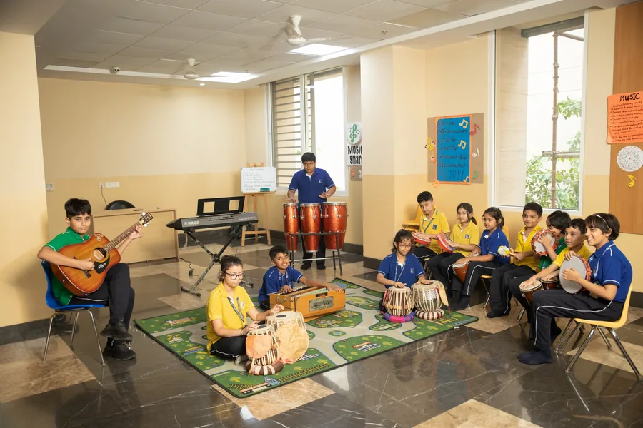 A teacher playing guitar while pupils play tabla and percussion on a patterned rug
