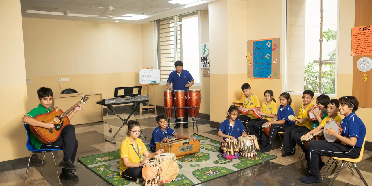 A teacher playing guitar while pupils play tabla and percussion on a patterned rug