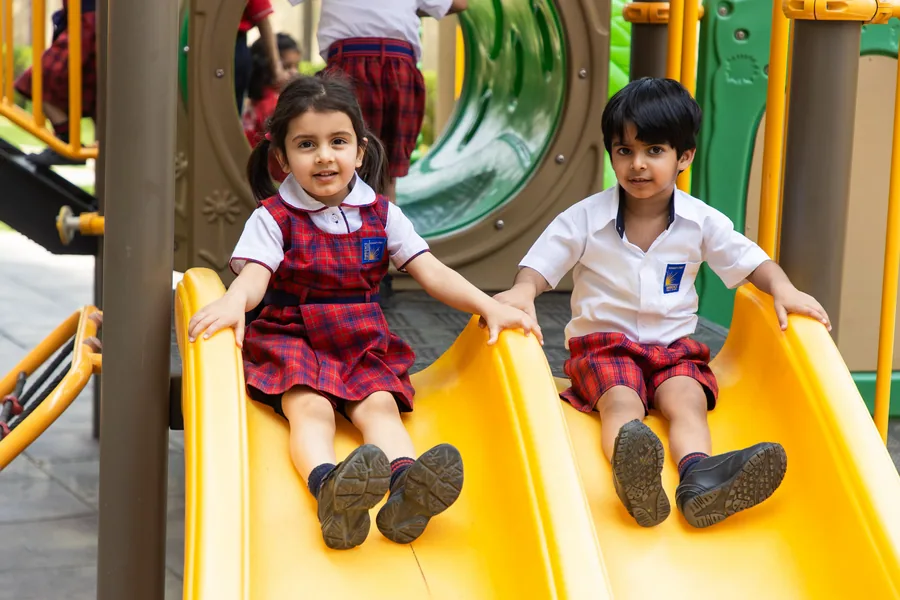 Two nursery children in red-check uniform coming down a yellow slide, both beaming