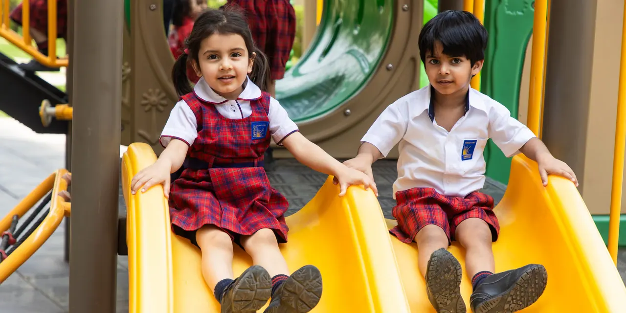 Two nursery children in red-check uniform coming down a yellow slide, both beaming